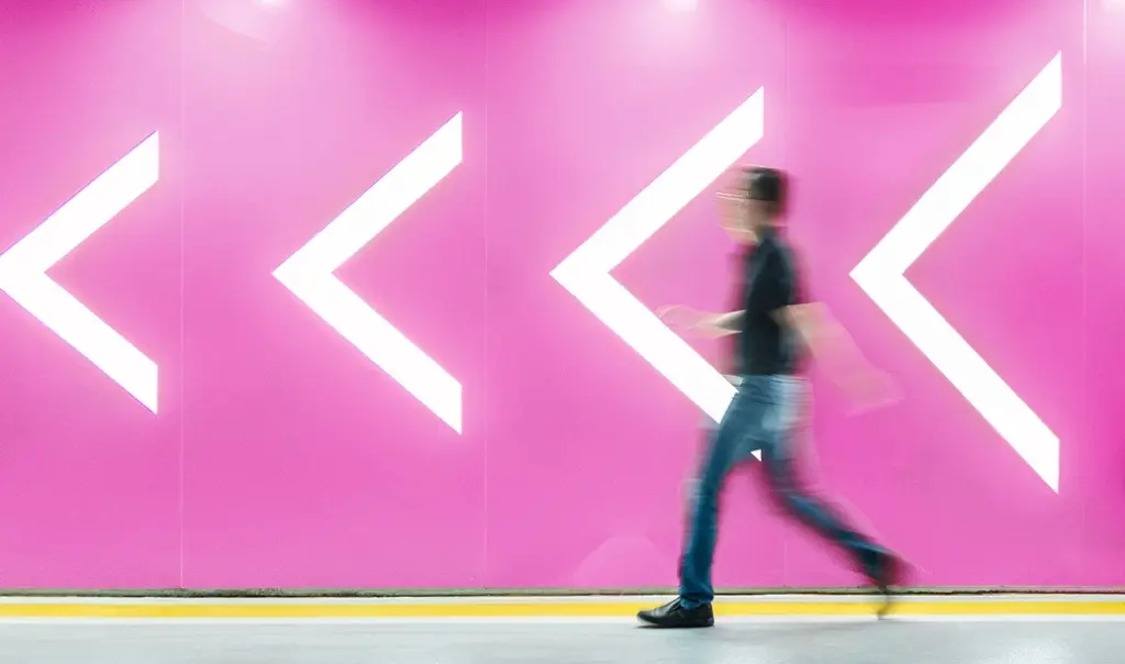 Man walking next to a pink wall with white arrows pointing left.